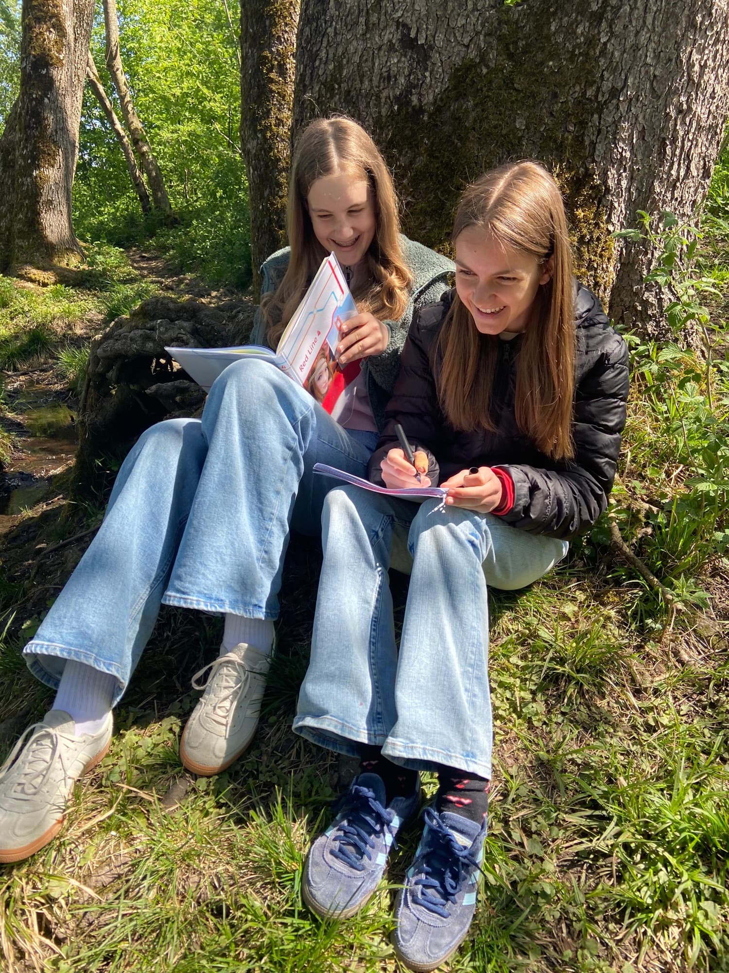 Zwei Kinder lernen gemeinsam mit Klemmbrett im Wald an einem Baumstamm.
